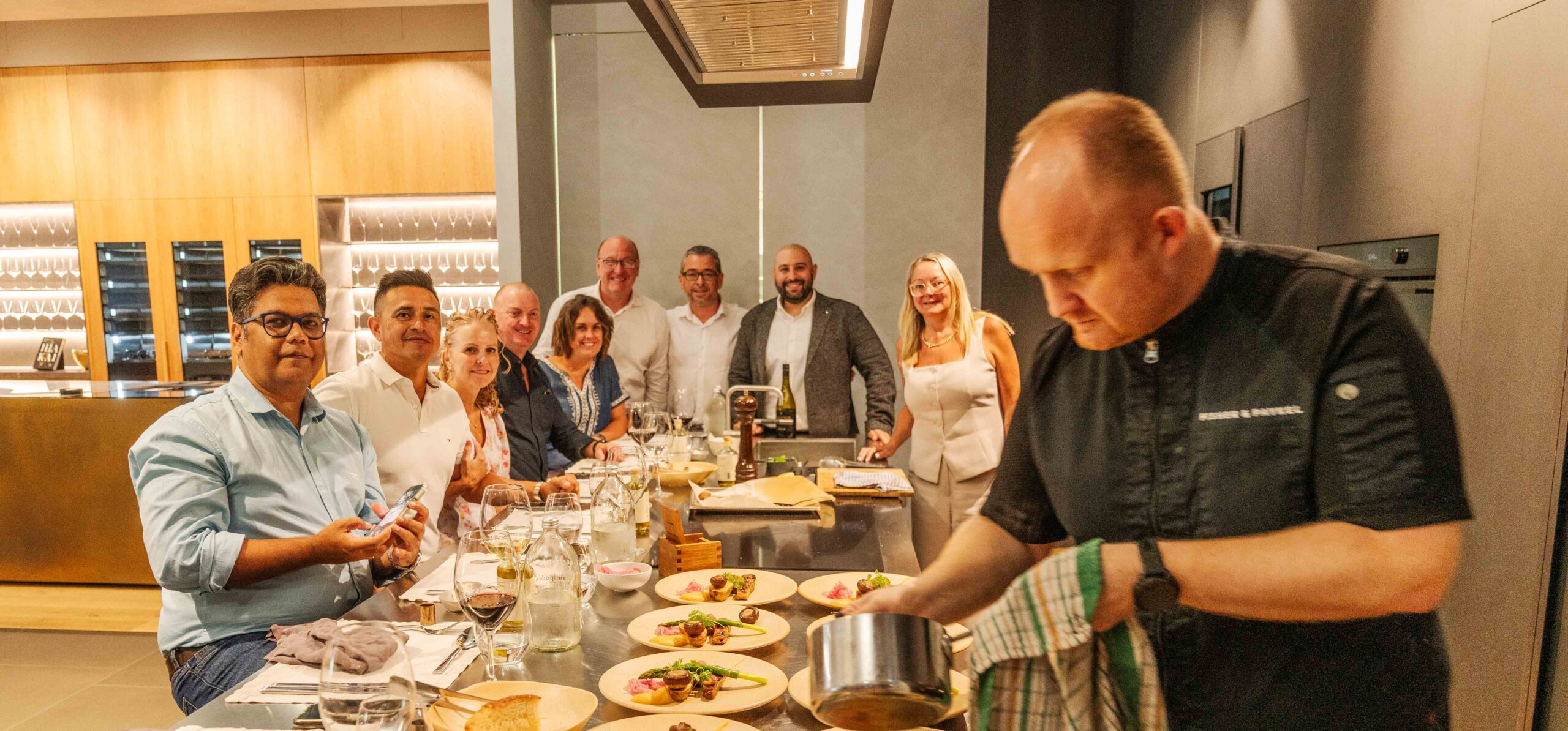 Chef Constantine, in his chef's whites, presents a beautifully plated dish to a group of smiling customers at the Fisher & Paykel Experience Centre.